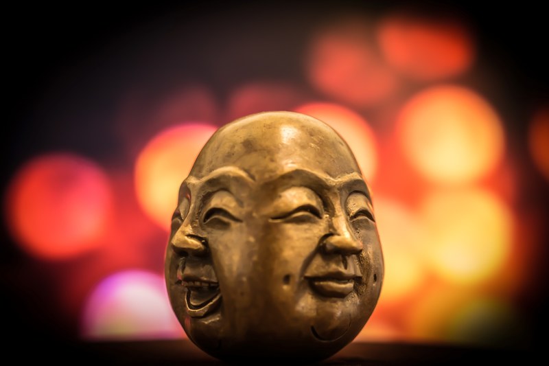 a brass figurine depicting two smiling faces of buddha with blurry colored lights in the background, two faces of buddha 4k