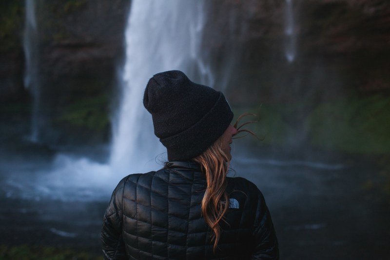 a woman in black winter attire is smiling and enjoying the scenery at seljalandsfoss waterfall, _woman seljalandsfoss waterfall 4k