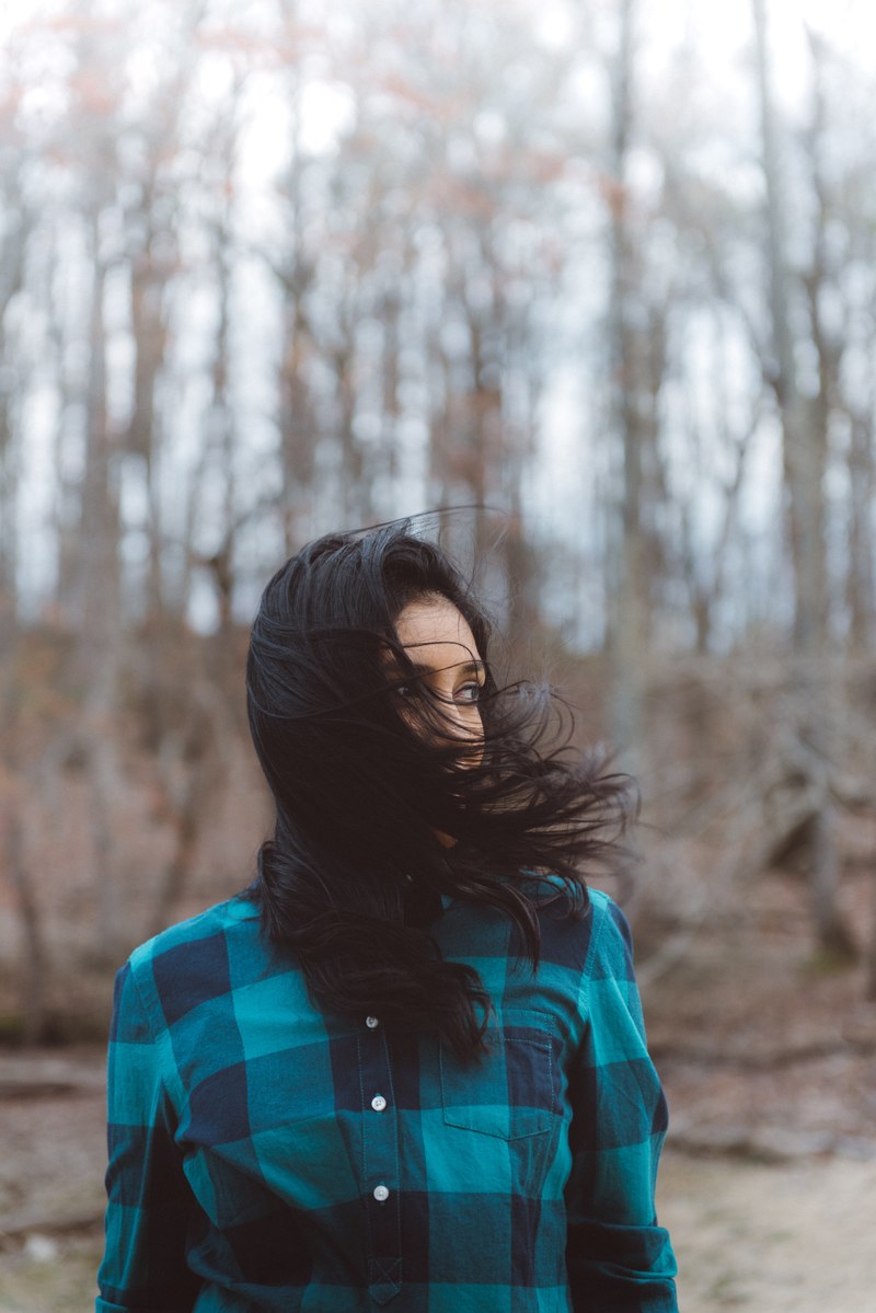 woman with black hair wearing a blue plaid flannel shirt in the woods, hair in her face 4k