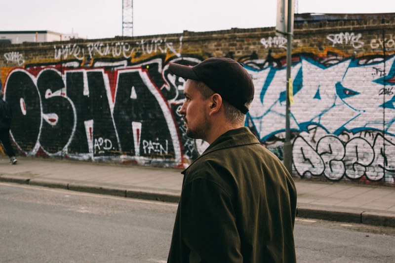 man wearing cap and jacket walks near hackney wick and passes graffiti, man walks in front of graffiti wall 4k