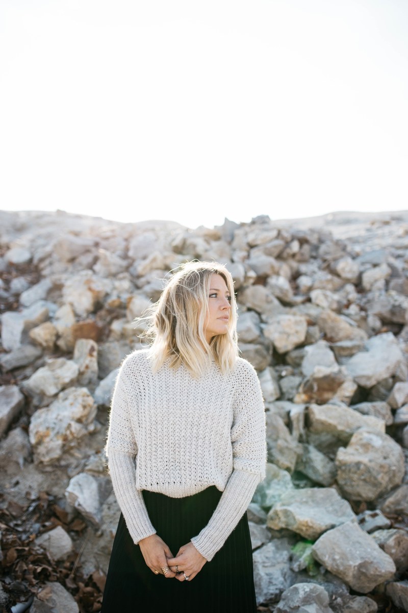 a young blonde woman in a white sweater standing near a large pile of stones, blonde woman by pile of stones 4k