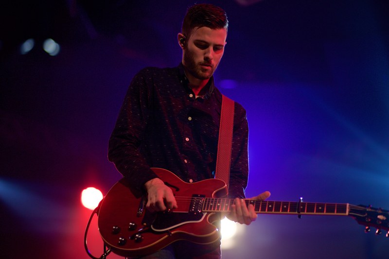 a male guitarist in a shirt performing on stage with blue and red lights behind him, guitarist on stage with blue and red lights 4k