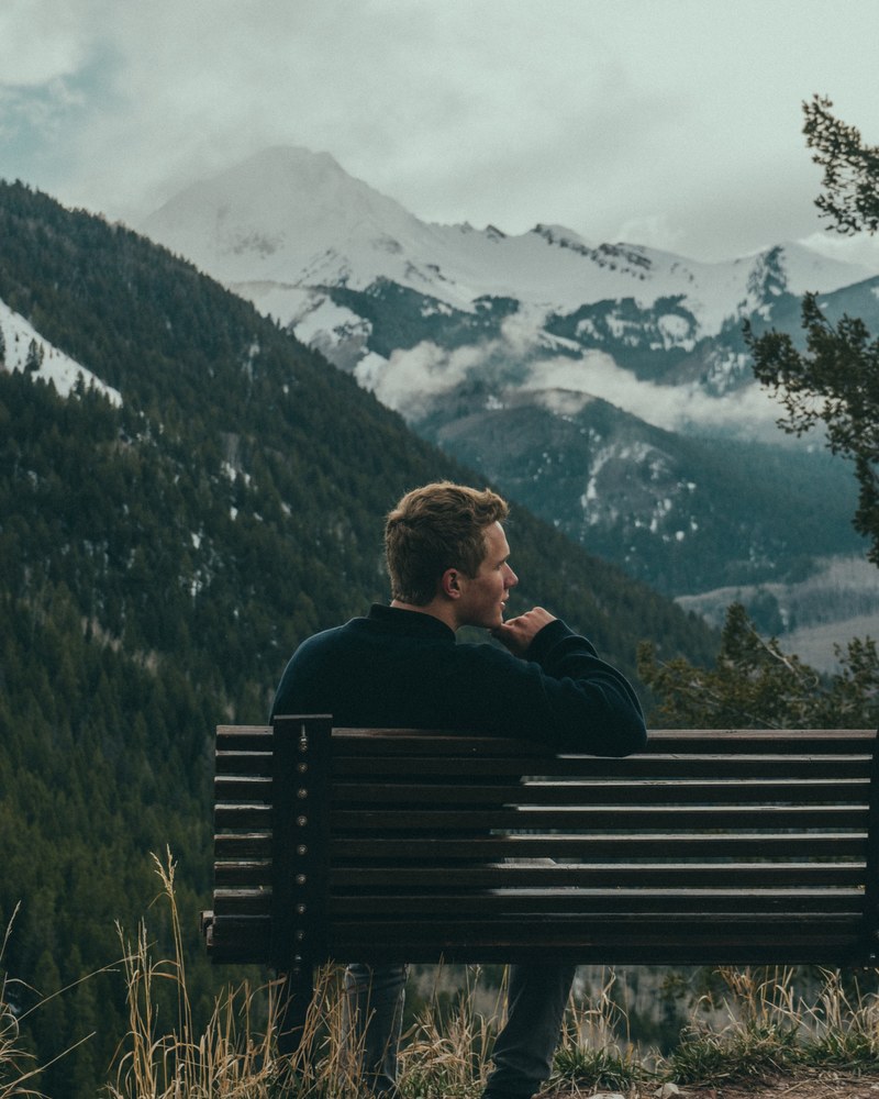 a young man sitting on a wooden bench overlooking wooded mountain slopes, bench in the mountains 4k