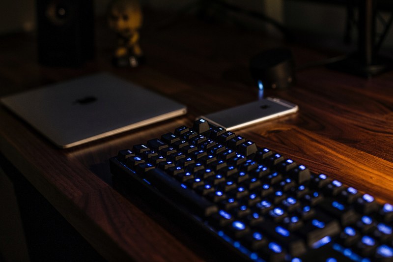 a dim picture of a blue backlit keyboard a smartphone and a tablet on a wooden desktop, battle amp workstation peripherals 4k