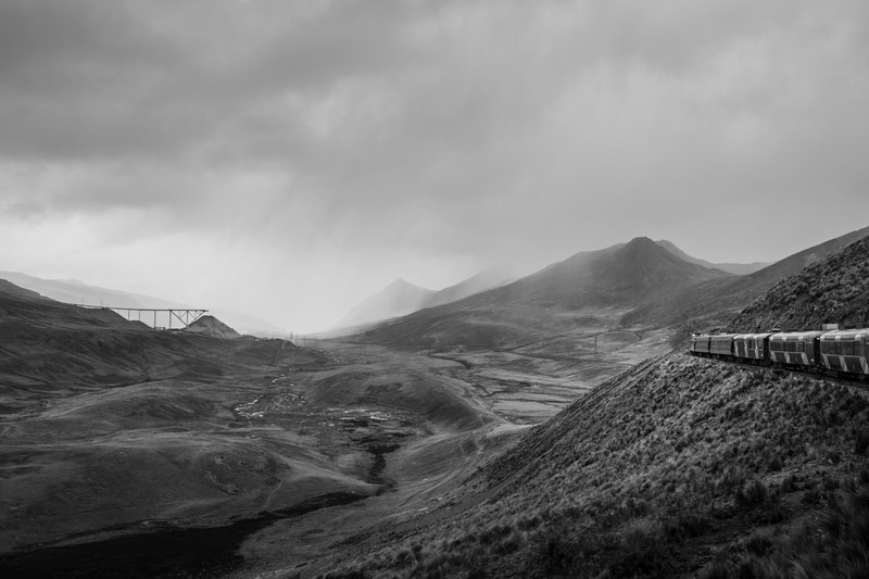 black and white panoramic shot of train travelling through mountains and rocks with cloudy sky, mountainous train ride 4k