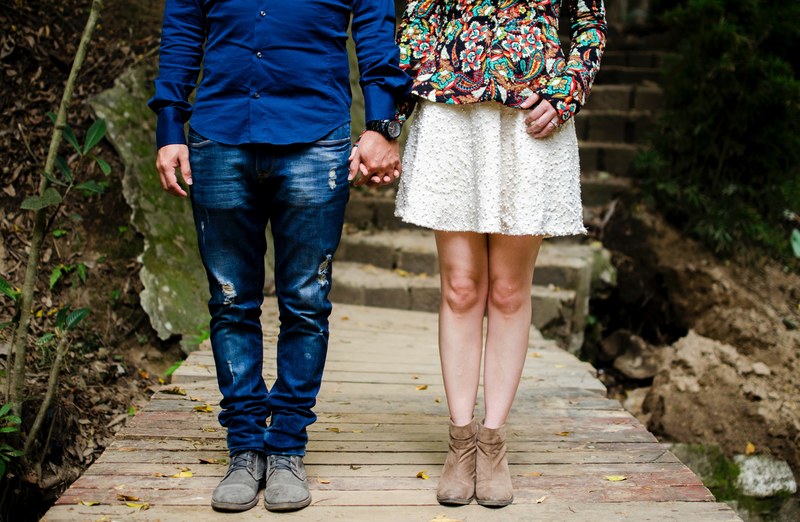 a couple in brightly colored clothes stands on a wood deck below stone stairs holding hands, holding hands on deck 4k