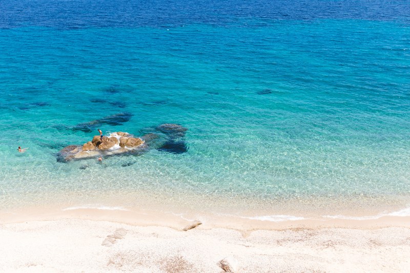 drone view of a person standing on a rock in a clear blue shallow ocean by the fava sand beach, blue ocean by the sand beach 4k