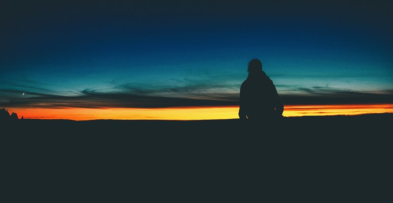 silhouette person standing in foreground of a black orange and blue sunrise or sunset horizon in montana, silhouette person standing with horizon 4k