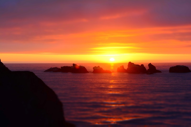 the sun setting on the horizon of the ocean at bandon with silhouetted rocks in the foreground, bandon sunset over the ocean and rocks 4k