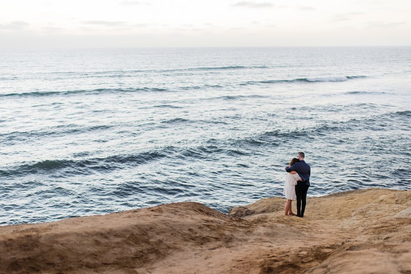 a couple stands on a rocky surface by the ocean their arms around each other, couple embrace sunset cliffs 4k