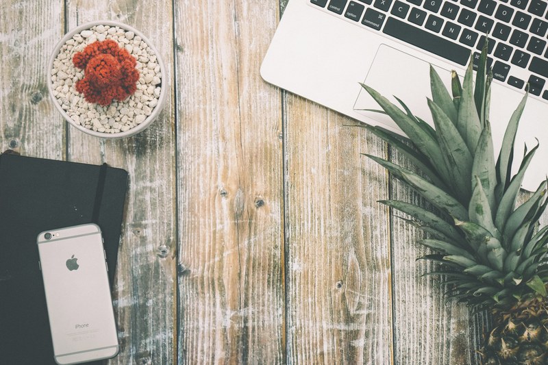 top view of a worn out wooden surface with a laptop a smartphone a notebook a red plant and a pineapple, tropical workspace 4k