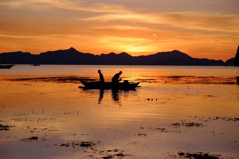 two fisherman in a boat their reflection on the water silhouetted against sun and orange sky, fishermen heading home after sunset 4k