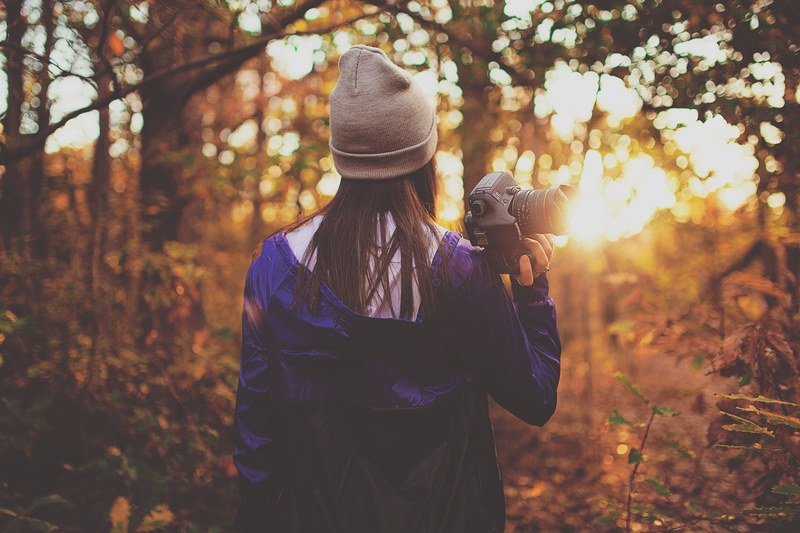 a woman in a beanie walking through a forest with a camera in her hand during sunset, photographer in woods at dusk 4k