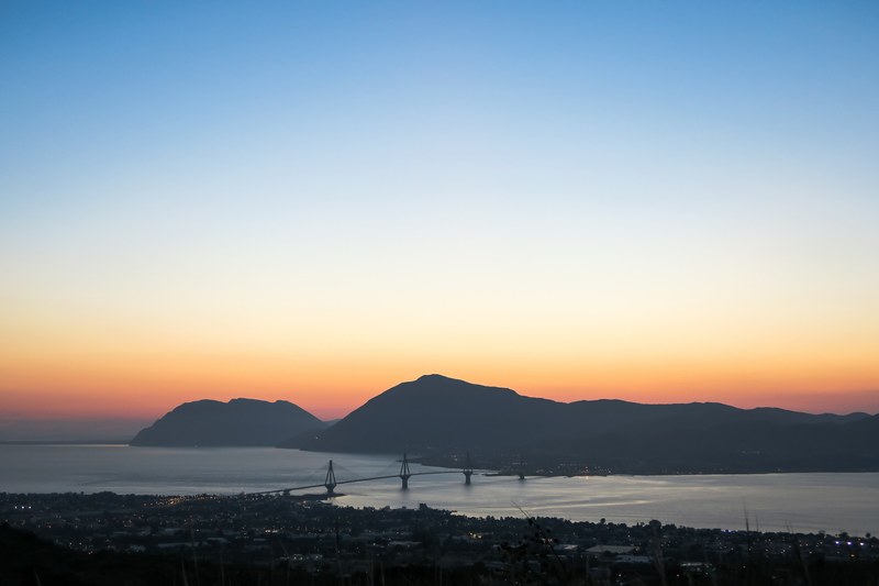 a sunset over a distant mountain and bridges over the river with the city of patras in the foreground, sunset over patras 4k