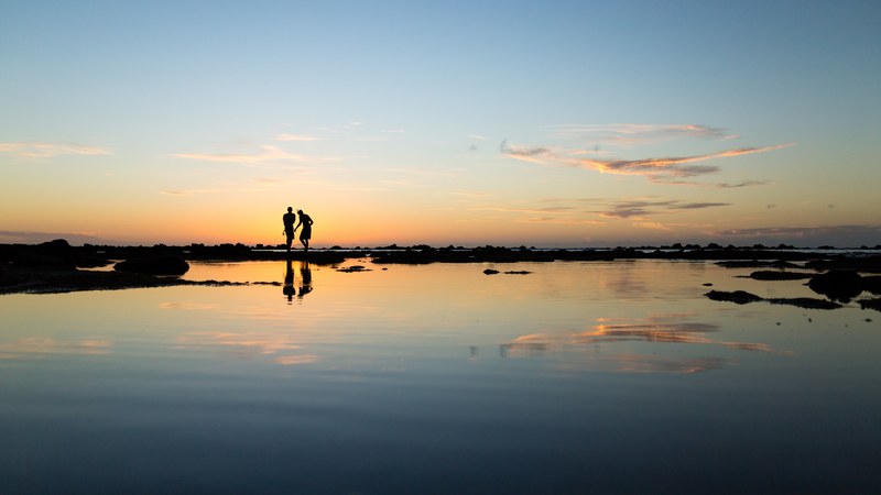 a silhouetted couple stand together on the horizon as they and the blue and yellow sky reflect into the still water foreground, two people at sunset 4k