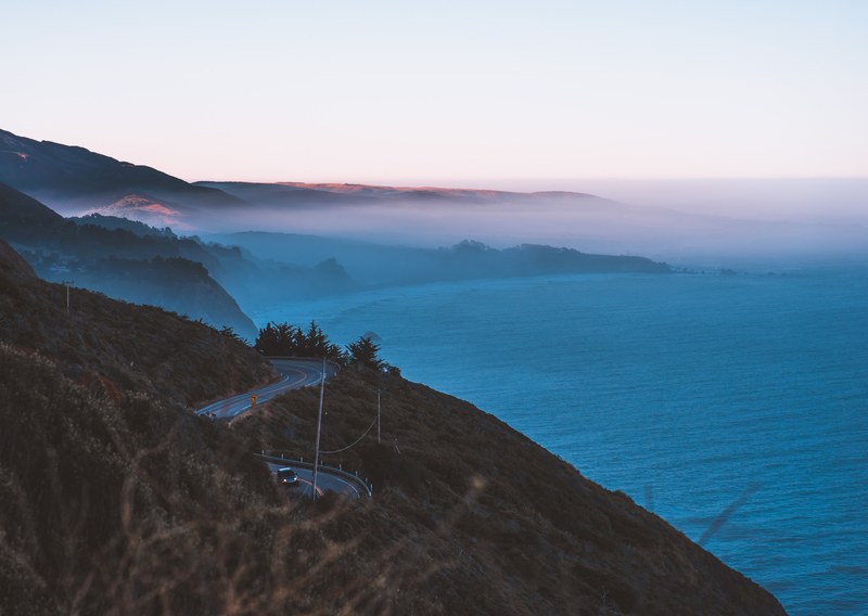 a road with cars winds around a mountain on the cost of the ocean, winding road near the seaside in big sur 4k