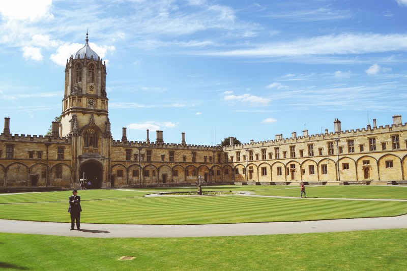 a person standing on a baseball field near a large stone campus in oxford, oxford college campus 4k