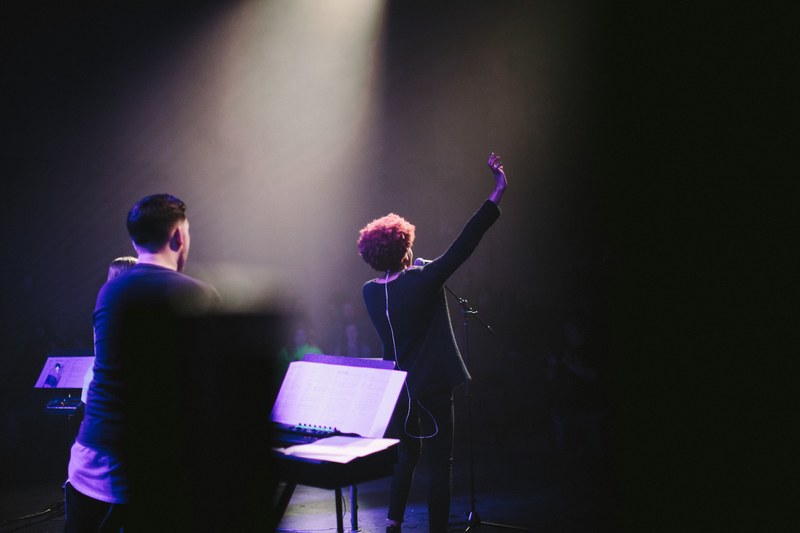 a woman at a microphone raises her hand on a dim stage, female vocalist waving 4k