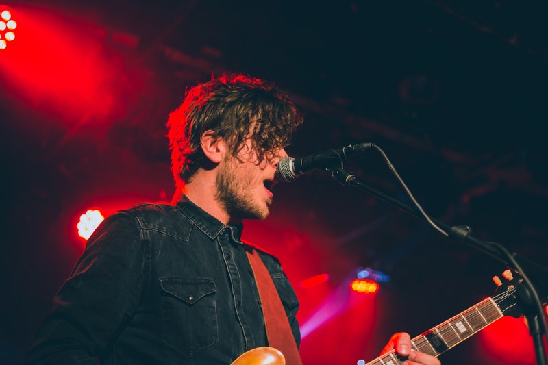 a bearded man with a guitar singing into a microphone with red lights behind his back, band member under red floodlights 4k