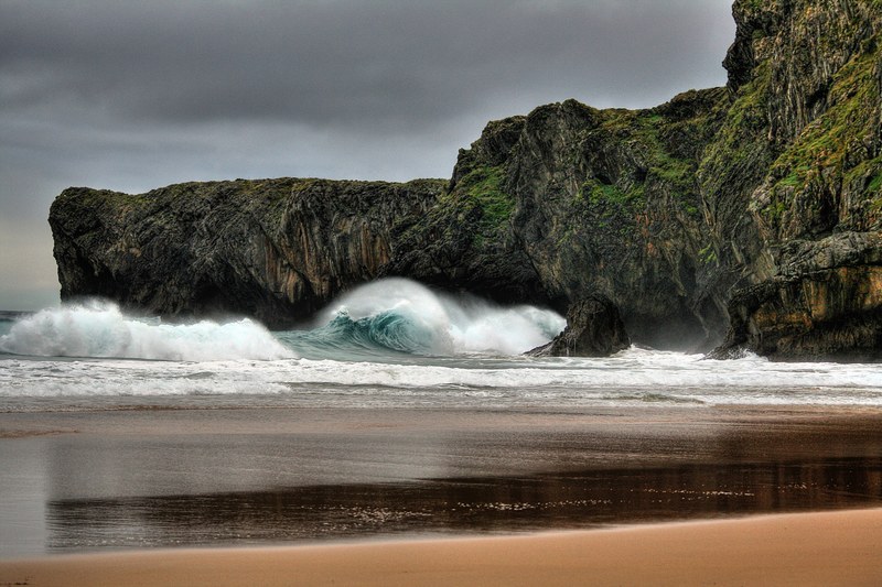 a rolling wave about to hit the rocky cliffs at andrn beneath a gray cloudy sky, rolling waves beneath the storm clouds 4k