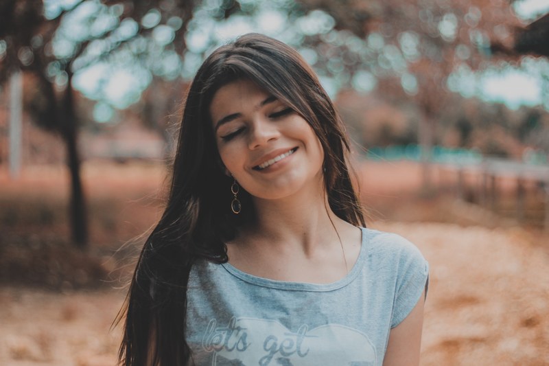a woman wearing long earrings smiles with closed eyes against a blurry outdoor background, closed eye smile 4k