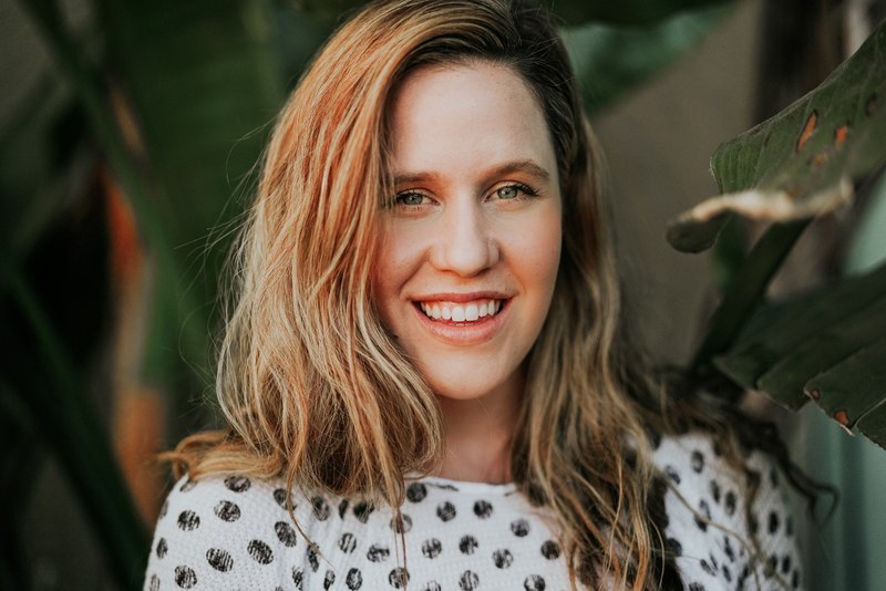 a blonde haired woman with a polka dotted shirt smiles surrounded by green plants, burnt sunshine 4k