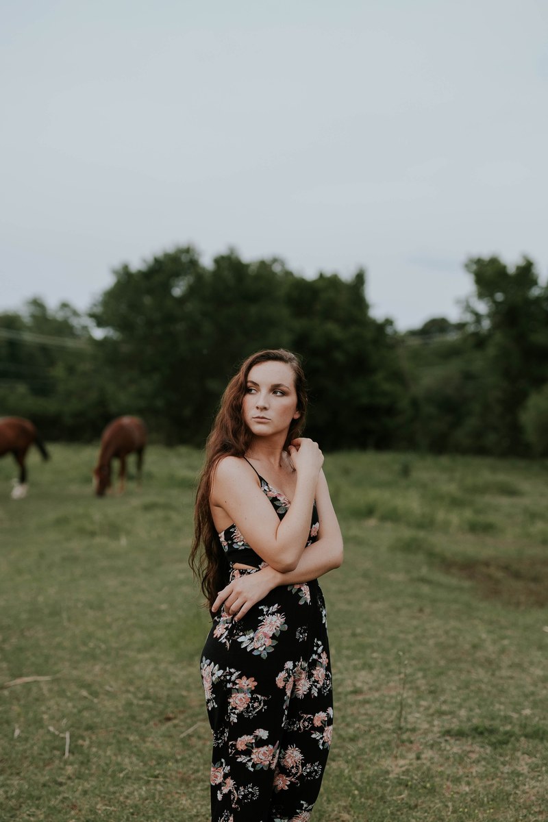 a young woman in a dress standing on a green pasture with horses grazing at the back, woman on a pasture 4k