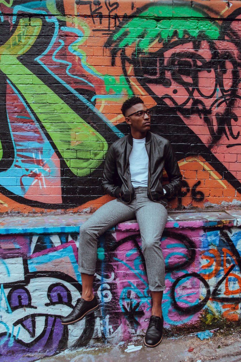 fashionable man in leather jacket sitting on wall in front of colorful graffiti leake street, urban fashion shot 4k