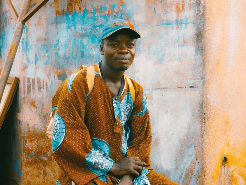 a man wearing a traditional orange and blue garment backpack and baseball hat near a graffitied wall, man in traditional outfit 4k