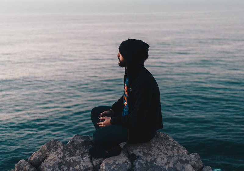 a male holding a camera and wearing a hoodie while sitting on a rock in figueira da foz looking out into the water, almost in the ocean 4k