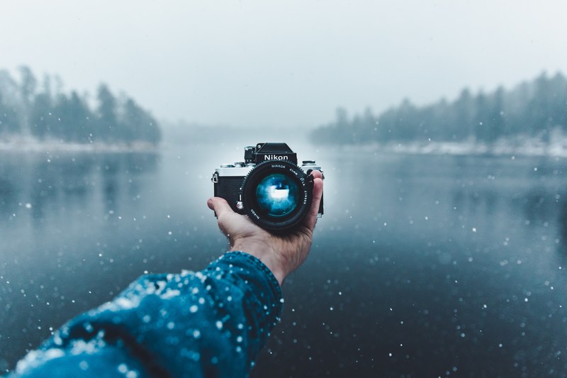photographer holding out antique nikon camera to take a picture of himself in snowy forest lake, nikon in the cold 4k