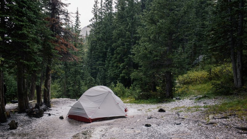 a single tent pitched on a gravel surfact amidst a wooded area with bright green trees, tent in a hail storm 4k