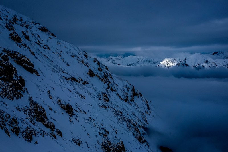 a blue hued shot of a snowy mountain side surrounded by thick clouds in the evening, blue clouds by the mountain side 4k
