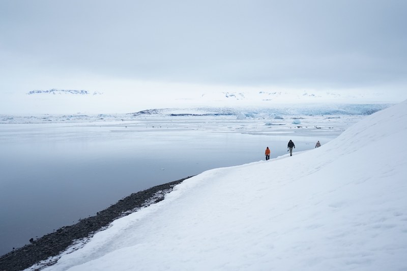 people walking along the bottom of a snowy mountain near an ocean, walking winter mountains 4k