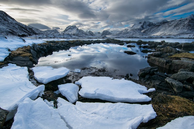 snow patches along rocks around a lake near mountains with cloud reflections in the water, bottom of mountain snow pond 4k