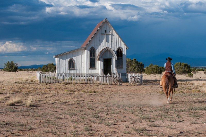 a man in a cowboy hat riding a horse near a wooden chapel in the middle of arid plains, cowboy at a desolate chapel 4k