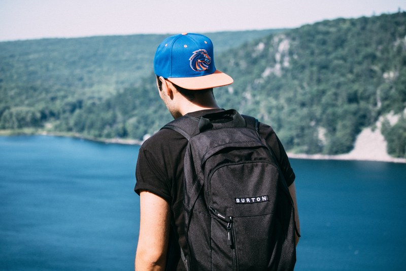the back of a man wearing a backpack and cap backwards looking out in devils lake by forest of trees, man looking out to lake 4k