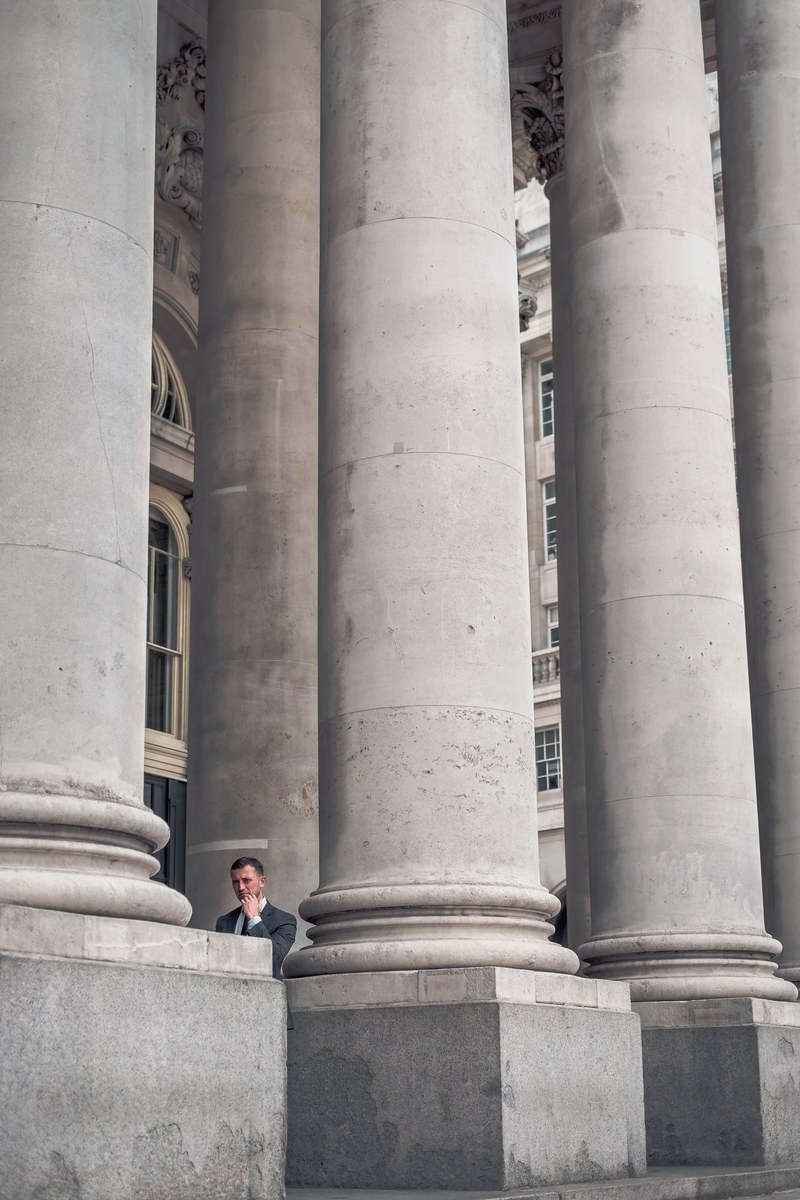 a man in a suit standing near the tall columns at the entrance to the london stock exchange, between the columns 4k