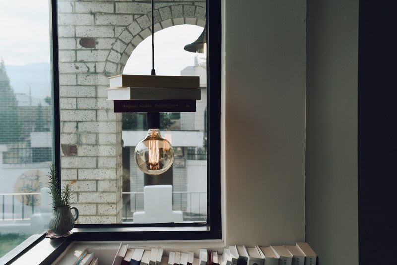lightbulb hangs from suspended books in a room with windows and a white brick building outside, lightbulb hangs from books 4k