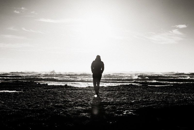 black and white shot of person walking towards sea water on shore with clouds in sky, person walks towards water 4k