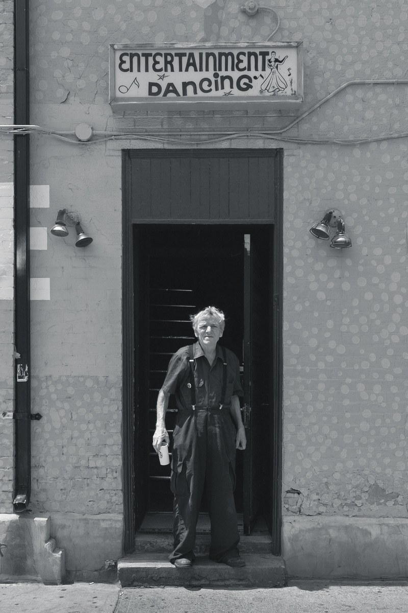 black and white shot of older man in dungarees holding drink in doorway with dancing sign, old man in doorway with drink 4k