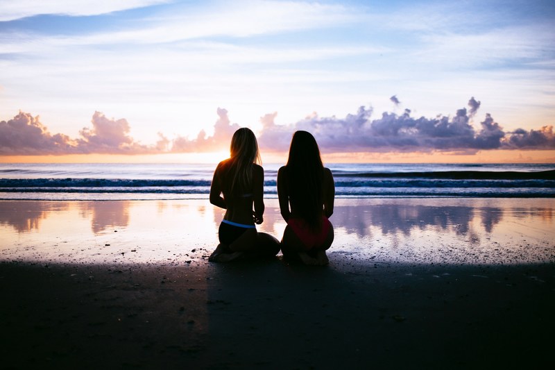 a dim shot of two women in bikini kneeling on a wet beach and admiring the sunset, watching the sunset on wet sand 4k