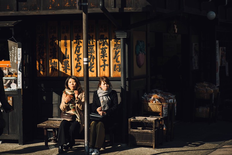 two women sitting in front of a wooden food store in japan and eating some snacks, two by two 4k