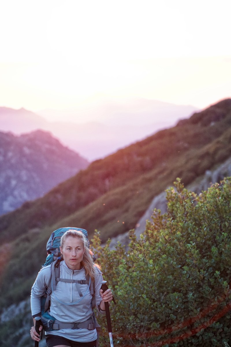 a blonde woman wears a backpack and uses walking poles while hiking near bushes a sunset over mountains behind her, sunrise_gr20_02 4k