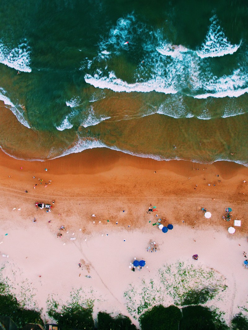 an aerial shot of waves breaking on a wet golden beach with tourists their umbrellas and deck chairs, top view of waves on a beach 4k
