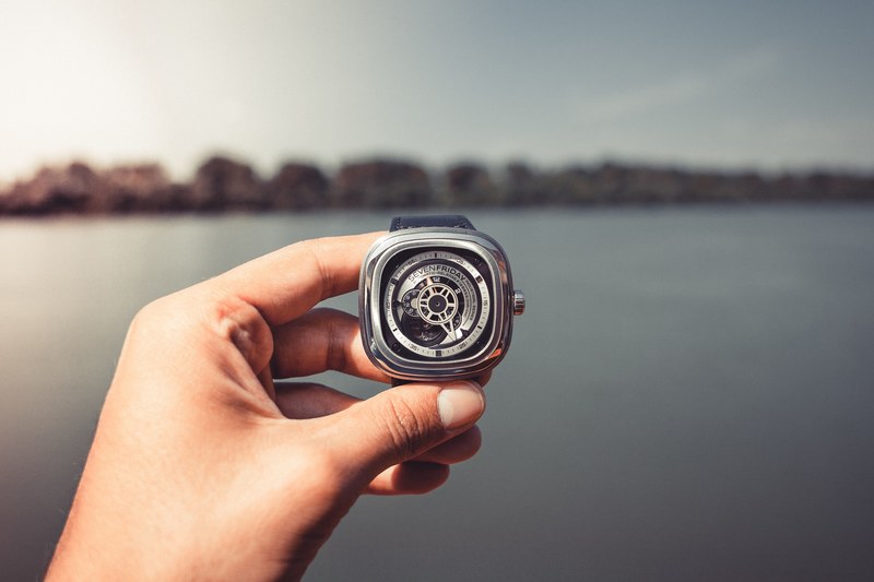 hand holding a modern watch with gears showing in front of a city skyline and water, sevenfriday watch 4k