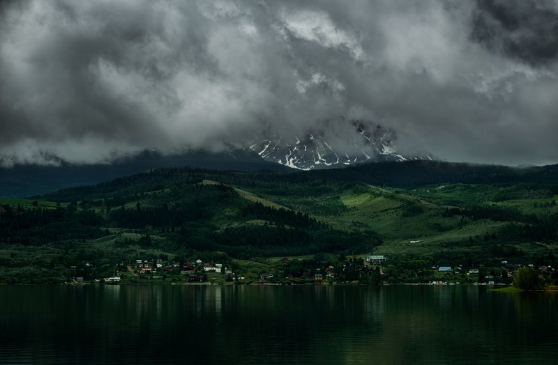 a village on a lake near tall mountains behind gray clouds, storm brewing over a village 4k