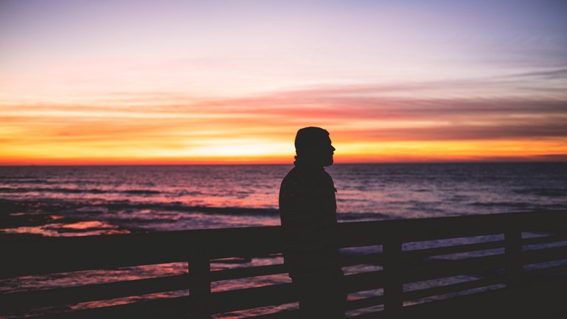 silhouette of a man looking into the distance while leaning against a fence by the ocean at dusk, walk on the pier 4k