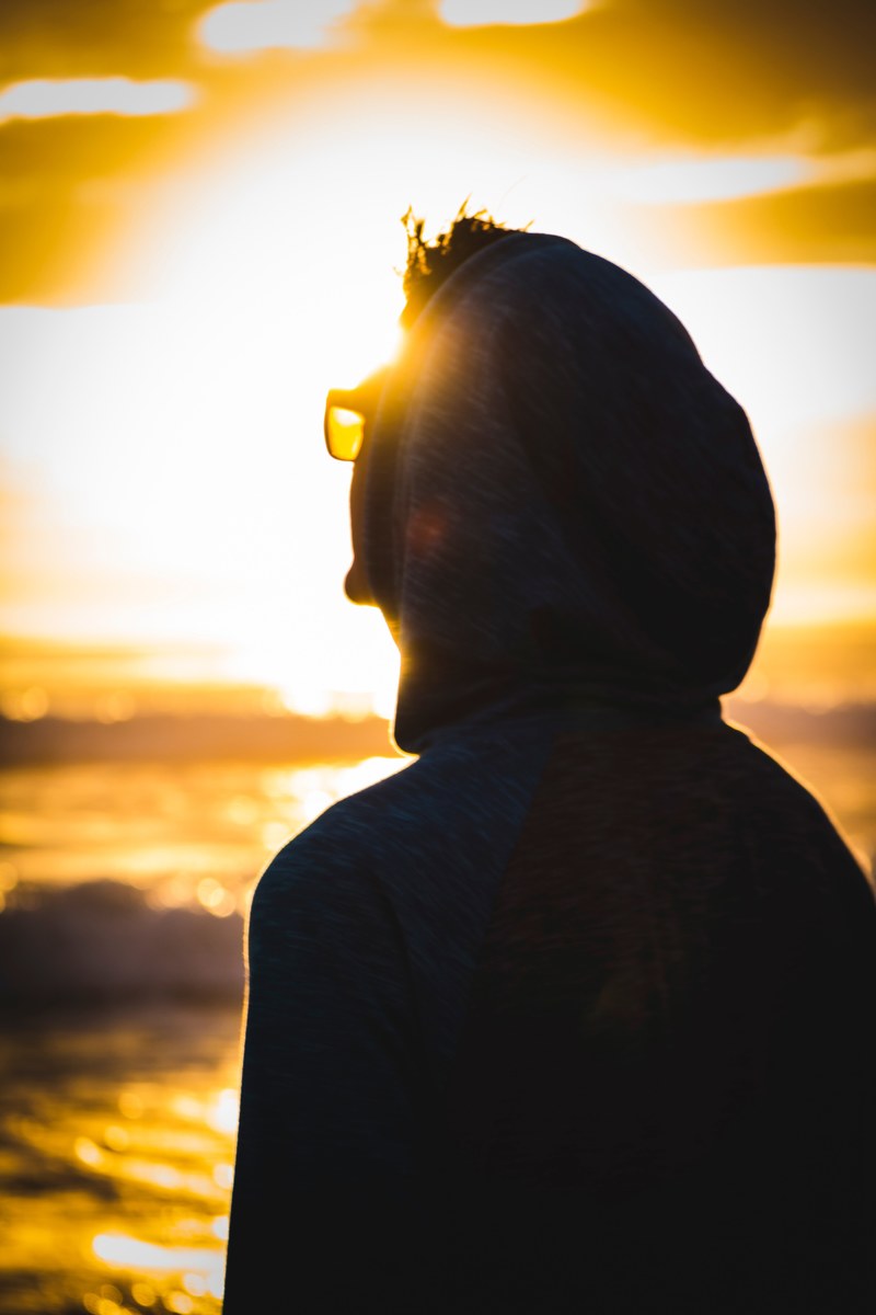 a person in glasses and a hoodie looks toward the bright sun in malibu, person with malibu sun 4k