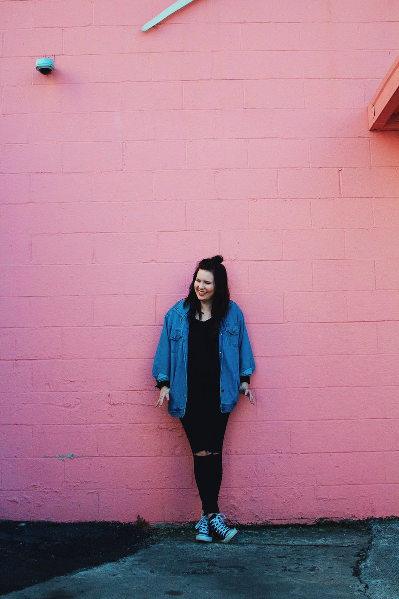 a smiling woman in a denim jacket and ripped jeans leaning against a pink wall, woman in ripped jeans 4k